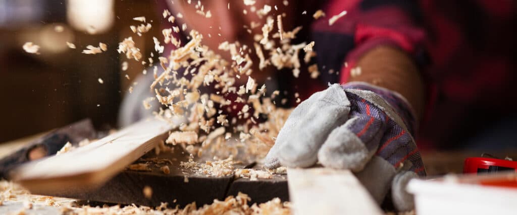 Artisan menuisier travaillant le bois avec des copeaux projetés dans l'air, portant un gant de protection.