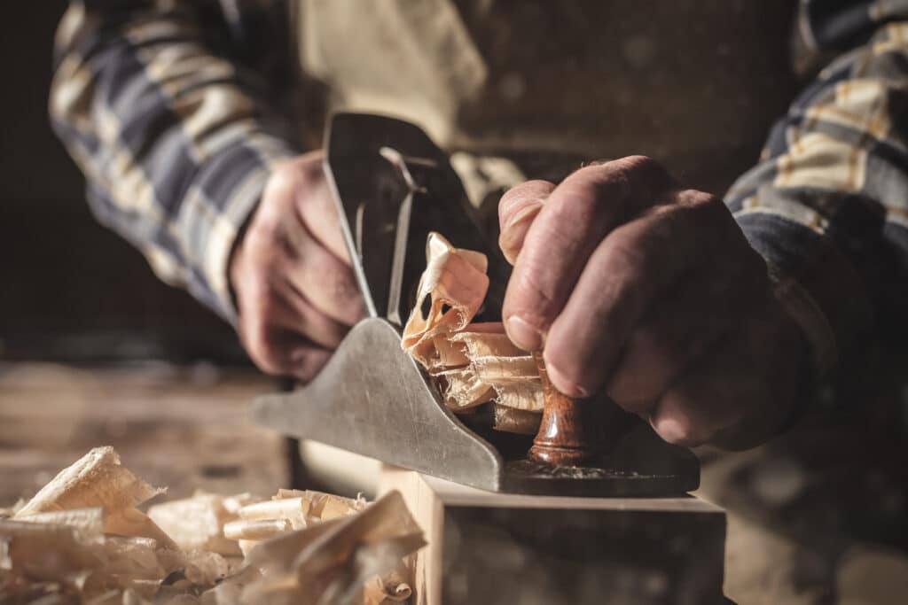 Artisan menuisier utilisant un rabot manuel sur du bois massif, projetant des copeaux fins.